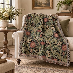 May include: A woven throw blanket featuring a dark background with a repeating botanical pattern of green artichokes, leafy foliage, and muted red flowers. The rectangular textile has a decorative fringed edge along the sides and bottom.