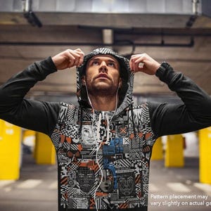 May include: A black long-sleeve hoodie featuring a complex, multi-colored graphic print on the front panel. The design includes abstract geometric shapes, digital patterns, and stylized facial outlines in shades of orange, blue, white, and black.