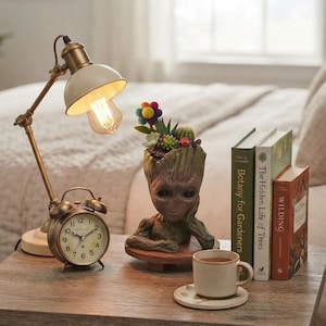 Op de afbeelding: Een bureaulamp van messing en crème, een ronde analoge wekker, een boomachtige plantenbak met kleine vetplanten en een viltbloem, een keramische mok op een schoteltje en drie boeken getiteld Botany for Gardeners, The Hidden Life of Trees en Wilding.