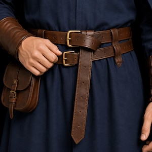May include: A dark brown leather double belt system worn over a navy blue garment. The belts feature brass-toned buckles and a matching leather pouch attached to the side. The leather has a smooth finish with visible stitching along the edges.