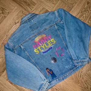May include: Light blue denim jacket featuring sequined text reading "Harry Styles" and various sequined patches. The jacket is hanging on a hanger. The background is wooden. The jacket has a classic collar and button closures.