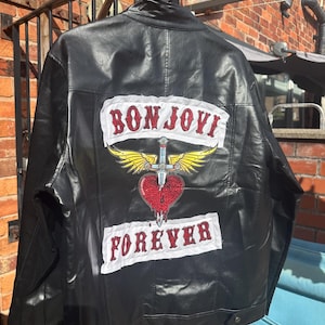 May include: A black jacket featuring a large back patch with the text BON JOVI in red rhinestones above a graphic of a winged dagger piercing a red rhinestone heart, with the word FOREVER in red rhinestones on a white patch at the bottom.
