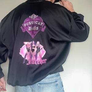 May include: A black satin bomber jacket featuring a purple heart graphic with the text Pussycat Dolls, a printed image of three performers in black outfits against a pink background, and the word Forever in pink script lettering on the back.