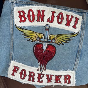 May include: A light blue denim jacket back panel featuring two white fabric patches with frayed edges. The top patch reads BON JOVI in red glitter letters. Below is a graphic of a winged dagger piercing a red glitter heart, with FOREVER in red glitter letters on the bottom patch.