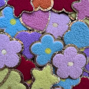 May include: A collection of assorted fabric patches in various shapes including flowers, hearts, and stars. The patches feature a textured, fuzzy surface in vibrant colors like pink, blue, orange, yellow, and red, all outlined with a gold glitter border.