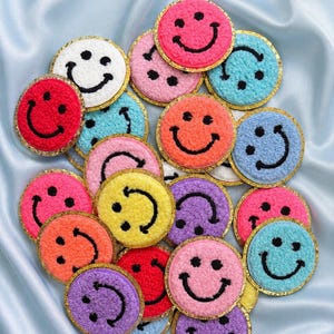 May include: A collection of circular patches featuring fuzzy, textured smiley faces in various bright colors including pink, blue, yellow, orange, purple, red, and white. Each patch is outlined with a thin, shimmering gold metallic border.