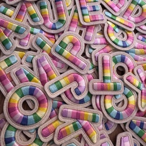 Puede incluir: Una colección de parches bordados con letras del alfabeto que presentan un vibrante patrón de rayas multicolor en tonos rosa, amarillo, verde y azul. Cada letra está delineada con un borde beige claro, creando un aspecto texturizado y superpuesto.