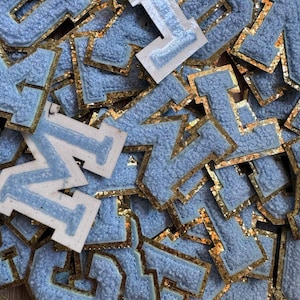 May include: A collection of light blue chenille letter patches featuring a gold glitter border. Each patch has a textured, fuzzy surface and a white felt backing. The letters are arranged in a pile, showing various characters including M, I, and others, with visible stitching along the edges of the gold trim.