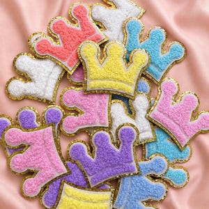 Può includere: Una collezione di toppe a forma di corona realizzate in materiale testurizzato e soffice in vari colori pastello, tra cui rosa, giallo, blu, viola e bianco. Ogni toppa a corona è delineata da un bordo glitterato color oro.