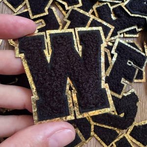 Op de afbeelding: Een verzameling letterpatches met een textuur van zwart pluizig materiaal in het midden en een rand van glinsterende gouden glitter. Elke patch heeft de vorm van een blokletter, waarbij een W wordt getoond om de details weer te geven.