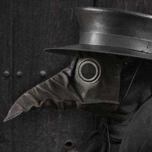 Op de afbeelding: Een zwart pestdoktermasker met een lange, snavelachtige neus en cirkelvormige oogopeningen. Het masker is gemaakt van leer en wordt gecombineerd met een hoge hoed met brede rand. Het masker is een historisch kostuum.