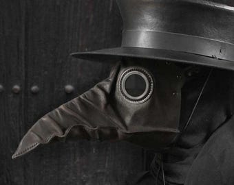 May include: A black plague doctor mask with a long, beak-like nose and circular eye openings. The mask is made of leather and is paired with a tall, wide-brimmed hat. The mask is a historical costume piece.