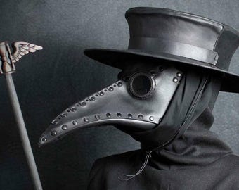 May include: A black leather plague doctor mask with a long, curved beak, round eye lenses, and a tall hat. The mask is part of a costume, and is paired with a black hooded cloak. A silver staff with a winged design is in the background.