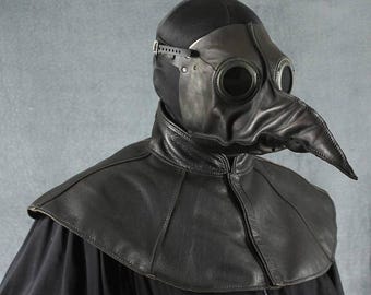 Può includere: Una maschera da medico della peste in pelle nera con un lungo becco e aperture oculari rotonde. La maschera ha un cappuccio nero ed è abbinata a un colletto in pelle coordinato. Il design ricorda gli abiti medici storici.
