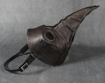Puede incluir: Una máscara de médico de la peste de cuero negro con un pico largo y curvado y grandes aberturas para los ojos. La máscara tiene una correa con una hebilla para sujetarla a la cara. La máscara está diseñada para ser usada.