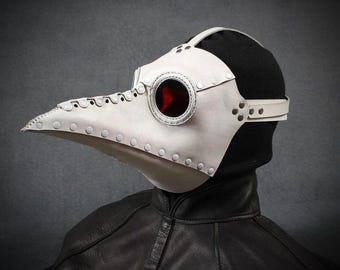 May include: A white leather plague doctor mask with a long, curved beak and a red lens. The mask is riveted and has a strap for securing it. The mask is worn over a black head covering and a black leather jacket.
