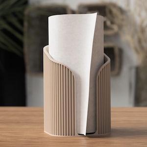 Pode incluir: Um porta-toalhas de papel bege com detalhes canelados verticais. O suporte tem uma abertura curva para dispensar toalhas de papel. Um rolo de toalhas de papel brancas é parcialmente visível dentro do suporte. O suporte está sobre uma superfície de madeira.