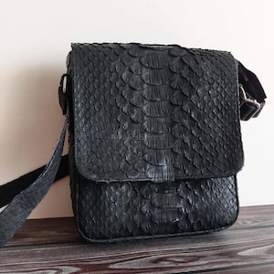 Könnte beinhalten: Schwarze Umhängetasche mit strukturierter, schuppenartiger Oberfläche. Die Tasche hat einen Klappverschluss, einen verstellbaren Riemen und eine Schnalle. Sie besteht aus dunklem Material und liegt auf einer Holzoberfläche.