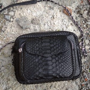 Op de afbeelding: Zwarte crossbodytas met een getextureerd, geschubd patroon, mogelijk slangenleer. De tas heeft een klepvak, een ritssluiting aan de bovenkant en een zilveren kettingriem. De tas is rechthoekig.