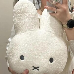 May include: A plush white bunny-shaped pillow with large, upright ears. The pillow has black dot eyes and a black 'X' for a mouth. The soft, textured material gives the pillow a cuddly appearance, perfect for a child's room or as a decorative item.