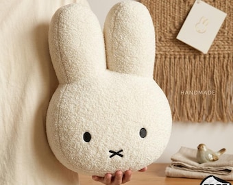 May include: A plush, off-white bunny-shaped pillow with black eyes and a stitched mouth. The pillow has a textured, fuzzy surface. The word "HANDMADE" is visible on a tag in the background. A small bird figurine and a folded cloth are also in the background.