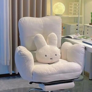May include: A white, plush office chair with a bunny-shaped pillow. The chair has a high back, armrests, and a five-star base with wheels. The bunny pillow has a simple face with black eyes and a cross for a mouth. The chair is in a well-lit room.