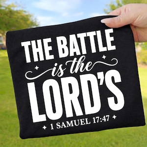 Könnte beinhalten: Ein schwarzes T-Shirt mit weißem Siebdrucktext, der THE BATTLE is the LORD'S und 1 SAMUEL 17:47 am unteren Rand zeigt. Das Design enthält dekorative Schnörkel und kleine vierzackige Sternakzente, die mittig auf der Vorderseite platziert sind.