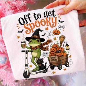 Könnte beinhalten: Ein weißes T-Shirt mit einem grünen Frosch, der einen Hexenhut und einen karierten Schal trägt, auf einem Roller fährt und einen Kaffee hält. Eine schwarze Katze und ein Holzwagen voller Kürbisse mit der Aufschrift Spooky Supplies sind abgebildet.