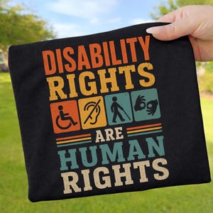May include: A black t-shirt featuring the text DISABILITY RIGHTS ARE HUMAN RIGHTS in a retro font. Between the lines are four square icons: a wheelchair user, a crossed-out ear, a person with a cane, and hands performing sign language.