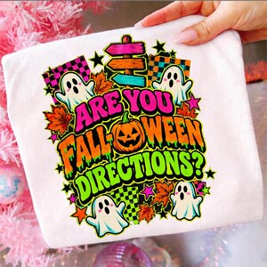 Könnte beinhalten: Ein weißes T-Shirt mit einer bunten Grafik und dem Text ARE YOU FALL-WEEN DIRECTIONS? Das Design zeigt drei weiße Geister, einen zentralen orangefarbenen Kürbis, Herbstblätter, Sterne, Karomuster und ein Wegweiser-Schild.