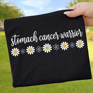 Könnte beinhalten: Ein schwarzes T-Shirt mit dem weißen, kursiven Schriftzug "stomach cancer warrior" auf der Brust. Unter dem Text befindet sich eine horizontale Reihe aus weißen Gänseblümchen mit gelber Mitte, durchsetzt mit kleineren weißen Blumenumrissen.