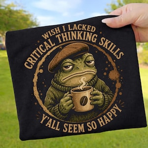 Könnte beinhalten: Ein schwarzes T-Shirt mit einer Grafik eines grünen Krötenwesens, das ein braunes Barett und einen grünen Pullover trägt und eine dampfende Tasse mit einem Kaffeebohnensymbol hält. Der Text lautet: WISH I LACKED CRITICAL THINKING SKILLS Y'ALL SEEM SO HAPPY.