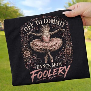Puede incluir: Camiseta negra con una rana bailarina en tutú rosa, con el texto "OFF TO COMMIT DANCE MOM FOOLERY" en una fuente decorativa. El diseño incluye detalles florales y está centrado en la camiseta.