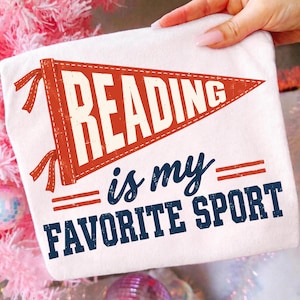 Könnte beinhalten: Ein weißes T-Shirt mit einer roten Wimpelgrafik, die das Wort READING in weißen Buchstaben zeigt. Unter dem Wimpel steht der Text is my FAVORITE SPORT in Dunkelblau, flankiert von zwei roten horizontalen Linien.
