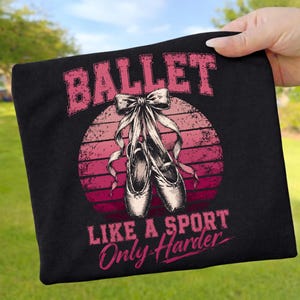 Könnte beinhalten: Schwarzes T-Shirt mit rosa Grafik. Das Wort "BALLET" steht über einem Paar Ballettschuhen mit Bändern. Darunter steht der Text "LIKE A SPORT Only Harder". Das Design zeigt einen rosa-roten Sonnenuntergang.