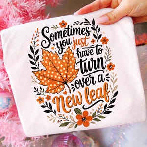 Könnte beinhalten: Ein weißes T-Shirt mit einem Grafikdesign, das ein großes orangefarbenes Ahornblatt mit weißen Punkten zeigt. Der Text lautet "Sometimes you just have to turn over a New leaf", umgeben von schwarzen Blattzweigen und kleinen orangefarbenen Blüten.