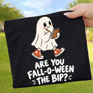 May include: A black t-shirt featuring a graphic of a white ghost wearing orange high-top trainers, holding a pencil and a notepad. Below the ghost, the white text reads: ARE YOU FALL-O-WEEN THE BIP? The design is centred on the front of the garment.