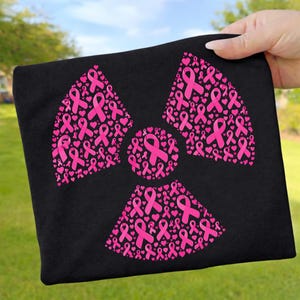 Könnte beinhalten: Ein schwarzes T-Shirt mit einem großen Strahlensymbol-Grafikdruck, der vollständig aus kleinen rosa Schleifen zur Brustkrebsvorsorge und winzigen Herzen besteht. Das Design ist mittig auf der Vorderseite des Kleidungsstücks platziert.