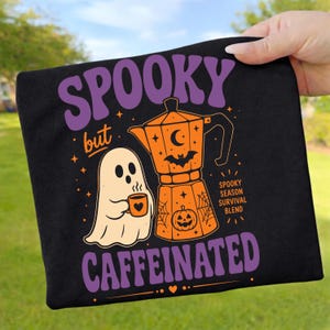 Könnte beinhalten: Ein schwarzes T-Shirt mit einem Aufdruck, der den violetten Schriftzug SPOOKY but CAFFEINATED zeigt. Das Motiv umfasst ein weißes Gespenst mit einer orangefarbenen Tasse, eine orangefarbene Espressokanne mit Fledermaus, Mond, Kürbis und Spinnenweben.