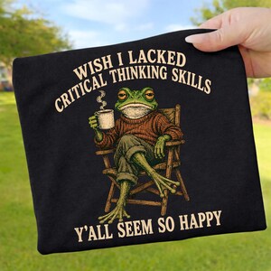 Puede incluir: Una camiseta negra con el gráfico de una rana verde con un suéter marrón, sentada en una silla de madera mientras sostiene una taza blanca humeante. El texto dice: WISH I LACKED CRITICAL THINKING SKILLS Y'ALL SEEM SO HAPPY en color beige.