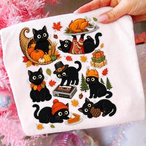 Könnte beinhalten: Ein weißes T-Shirt mit einem Grafikdruck von mehreren schwarzen Katzen in herbstlichen Szenen. Die Katzen sind mit Kürbissen, einem Füllhorn, einem gebratenen Truthahn, einem Kassettenrekorder, einem Stück Kürbiskuchen und Herbstblättern dargestellt.