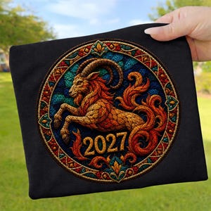 Könnte beinhalten: Ein schwarzes T-Shirt mit einer kreisförmigen Grafik im Buntglas-Stil, die eine Ziege mit geschwungenen Hörnern und einem fließenden Schwanz zeigt. Das Design enthält leuchtende Orange-, Rot-, Blau- und Grüntöne mit der Zahl 2027 unten.