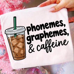 Pode incluir: Uma t-shirt branca com um gráfico de um café gelado num copo de plástico transparente com uma palhinha verde e cubos de gelo. À direita do copo, o texto preto diz: phonemes, graphemes, & caffeine. O design está impresso na frente da t-shirt.
