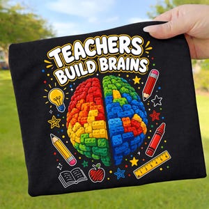 Könnte beinhalten: Schwarzes T-Shirt mit dem Text "TEACHERS BUILD BRAINS" über einer bunten Gehirngrafik. Das Gehirn ist in zwei Hälften geteilt, eine rot und gelb, die andere blau und grün, aus Bausteinen. Enthält Bleistifte, Sterne und ein Lineal.