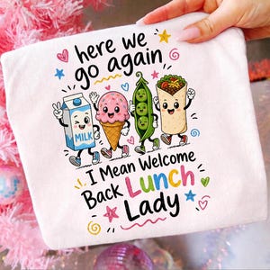 Könnte beinhalten: Ein weißes T-Shirt mit einer Grafik aus Cartoon-Figuren wie einem Milchkarton, einer Eistüte, einer Erbsenschote und einem Burrito mit Gesichtern. Der Text lautet "here we go again" und "I Mean Welcome Back Lunch Lady" mit bunten Sternen.