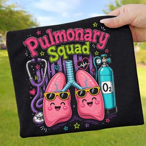 May include: A black t-shirt featuring a graphic with the text Pulmonary Squad in pink and green. The design includes a purple stethoscope, two pink lungs with smiling faces wearing black sunglasses, a heart with an EKG line, and a blue oxygen tank.