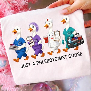 Könnte beinhalten: Ein weißes T-Shirt mit einer Grafik von vier Gänse-Figuren in medizinischer Kleidung. Die Gänse tragen Arztkittel, OP-Kleidung und eine Haube und halten Reagenzgläser sowie eine Checkliste. Der Text lautet JUST A PHLEBOTOMIST GOOSE.