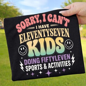 以下が含まれることがあります： 「SORRY, I CAN'T I HAVE ELEVENTYSEVEN KIDS DOING FIFTYLEVEN SPORTS & ACTIVITIES」とピンク、黄色、緑、白で書かれた黒いTシャツ。スマイリーフェイスと稲妻のグラフィックが含まれています。