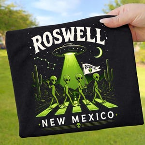 Könnte beinhalten: Schwarzes T-Shirt mit einem grafischen Design mit dem Text "ROSWELL NEW MEXICO". Das Design umfasst ein UFO, Aliens, die einen Zebrastreifen überqueren, Kakteen und eine Konstellation. Die Aliens halten eine Flagge mit der Aufschrift "ROSWELL".