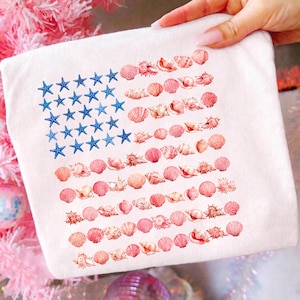 Puede incluir: Una camiseta blanca con un gráfico de la bandera estadounidense compuesto por estrellas de mar azules en el cantón y filas de varias conchas marinas de color rosa y melocotón que forman las rayas. El diseño está impreso en la tela.